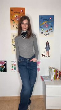 Ung kvinne i grå langermet skjorte og jeans står med hånden i lommen mot en vegg med fargerike plakater og en liten hvit kommode.