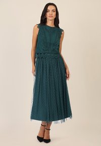 Tealfarbenes Spitzenkleid mit gewellten Kanten, eng anliegendem Oberteil und fließendem Tüllrock mit Pünktchen. Dazu schwarze Schuhe mit Knöchelriemen.