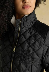 Čierna prešívaná bunda so zlatým zipsom, s semišovou záplatou na ramene a vyšívaným logom na hrudi. Dizajn s vysokým golierom.