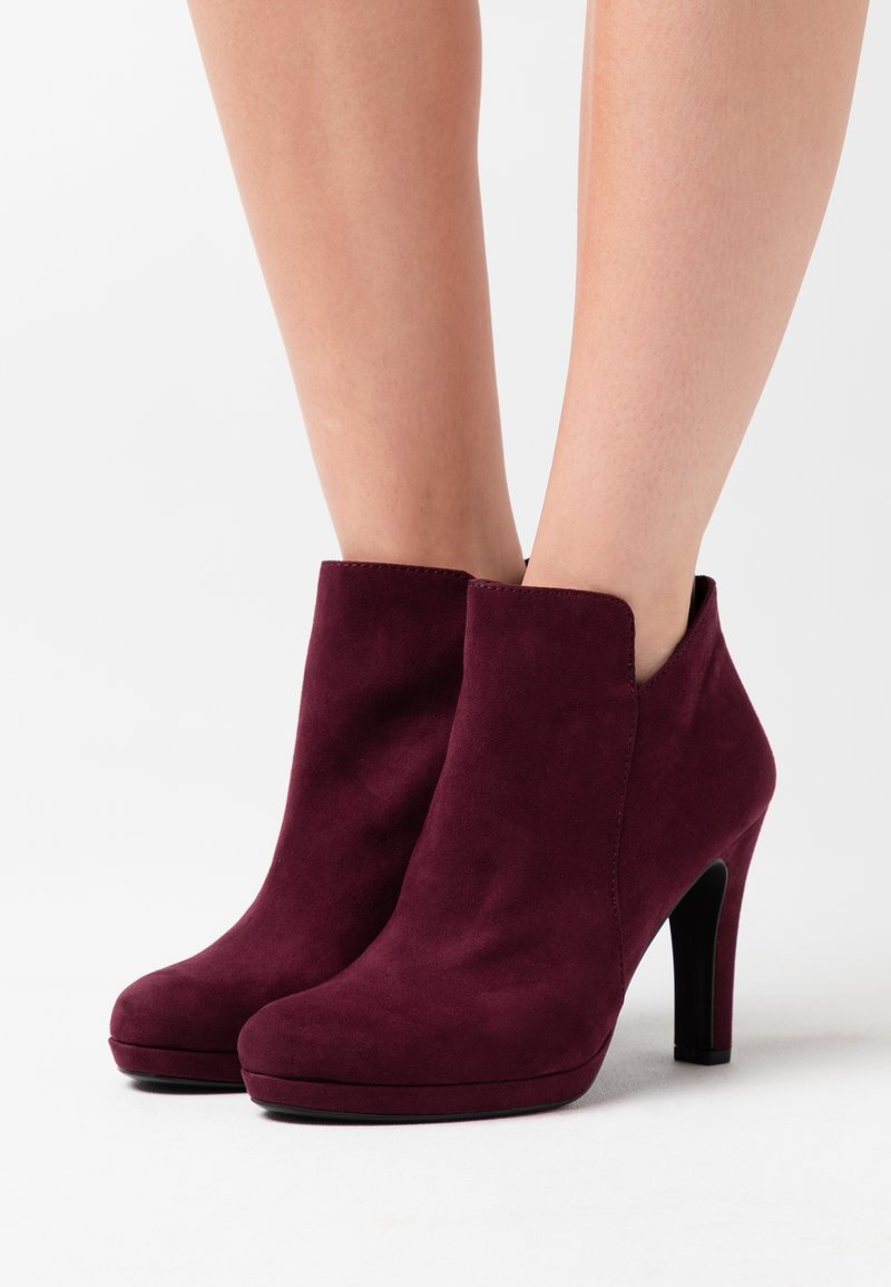 Femme portant des bottines en daim rouge foncé à talons hauts et légère plateforme, debout devant un fond clair uni.