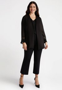 Giacca nera aperta davanti con maniche trasparenti e dettagli in pizzo, abbinata a un top nero e pantaloni cropped. Presenta tacchi a stiletto con punta a forma di freccia.