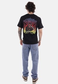 Uomo con tatuaggi, maglietta nera con grafica di pantera ruggente e testo viola, jeans azzurri e sneakers Nike marroni, visto di spalle.