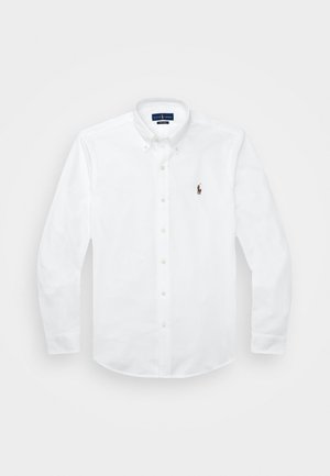 Chemise blanche à manches longues avec col boutonné et petit logo brodé sur la poitrine gauche, posée à plat sur fond blanc.