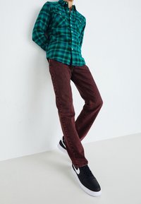 Persona apoyada contra la pared, vestida con una camisa de cuadros verde, jeans burdeos y zapatillas negras y blancas, posando con las piernas cruzadas.