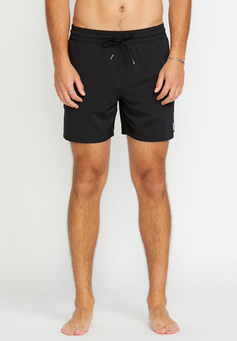 Torso et jambes d'un homme portant un short de bain noir à cordon, debout pieds nus sur un fond clair uni.