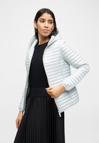 Γυναίκα που φοράει ανοιχτό γκρι καπιτονέ μπουφάν με κουκούλα πάνω από μαύρο τοπ και πλισέ μαύρη φούστα, κοιτάζει προς τα πλάγια και κρατάει το φερμουάρ του μπουφάν.