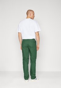 Pantaloni din bumbac verzi cu picior drept și buzunare la spate, purtați cu un tricou alb simplu și adidași albi.