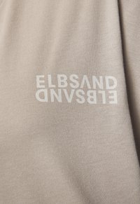 Beige Stoff T-Shirt mit auffälligem, inversen weißen Text "ELBSAND" über der Brust, mit einer glatten, leicht strukturierten Oberfläche.