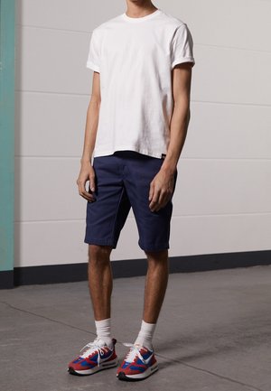 Homme debout portant un t-shirt blanc uni, un short bleu marine, des chaussettes blanches et des chaussures de sport rouges, bleues et blanches sur un sol gris.