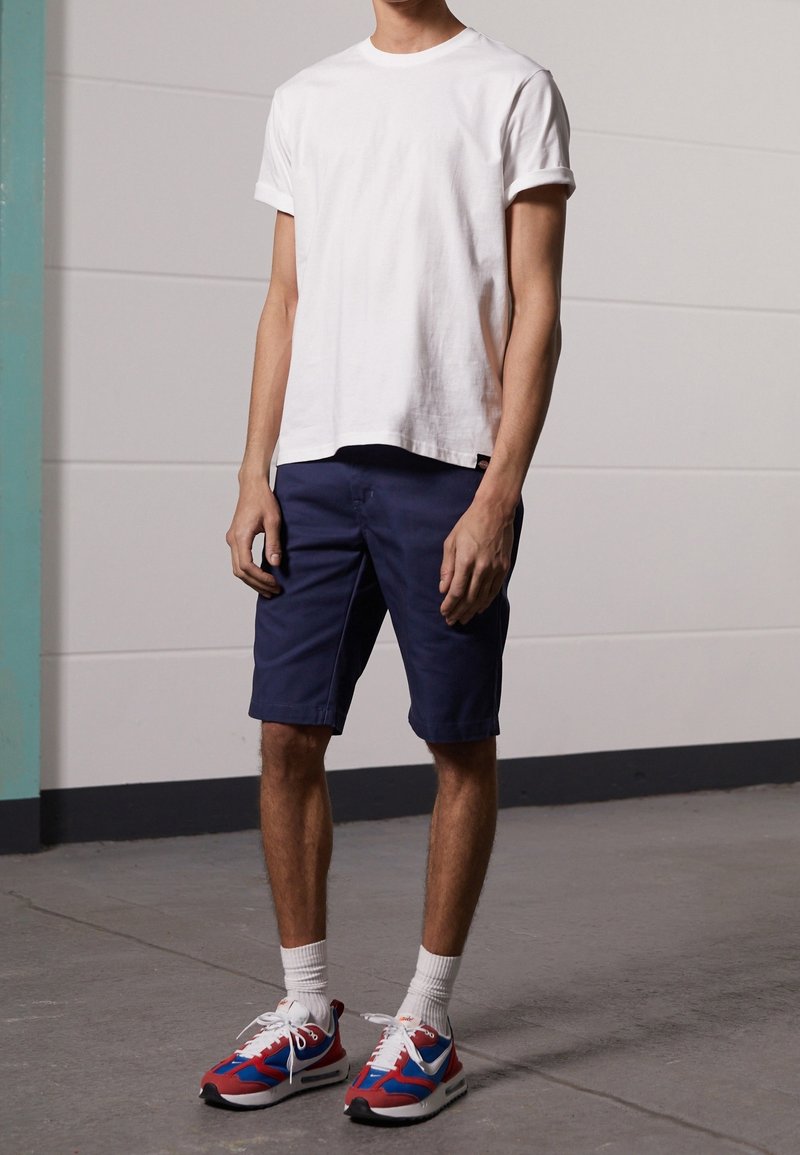 Homme debout portant un t-shirt blanc uni, un short bleu marine, des chaussettes blanches et des chaussures de sport rouges, bleues et blanches sur un sol gris.