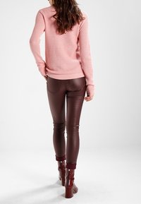 Ljusrosa strukturerad stickad tröja, matchad med bruna figurnära läderbyxor och vinröda klackade ankelstövlar. Bakvy, neutral bakgrund.