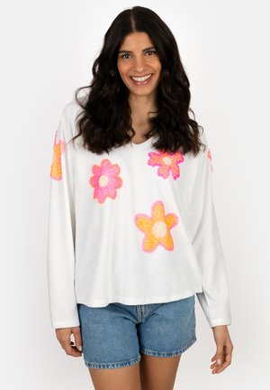 Lächelnde Frau mit langen dunklen Haaren, die ein weißes, langärmeliges Oberteil mit großen pinken und orangefarbenen Blumenmustern und Jeansshorts trägt.