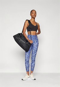 Fekete sportos duffel táska fehér logóval, kék mintás leggingssel és fekete crop toppal párosítva. A modell sportcipőben áll.