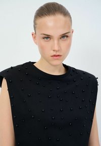 Top nero con una vestibilità ampia, decorato con perline nere in rilievo sulla parte frontale e un ampio scollo. Tessuto liscio e opaco.