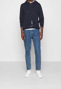 Man i marinblå hoodie med subtil logotyp, blå raka jeans och vita sneakers, stående mot en enkel vit bakgrund.