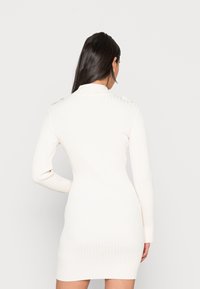 Abito bianco a costine con maniche lunghe, collo alto e una silhouette aderente. Caratterizzato da dettagli sulle spalle e una finitura testurizzata.