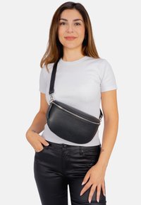 Schwarze Leder-Crossbody-Tasche mit strukturierter Oberfläche, runder Form, silbernem Reißverschluss und verstellbarem Trageriemen, getragen über einem weißen T-Shirt.