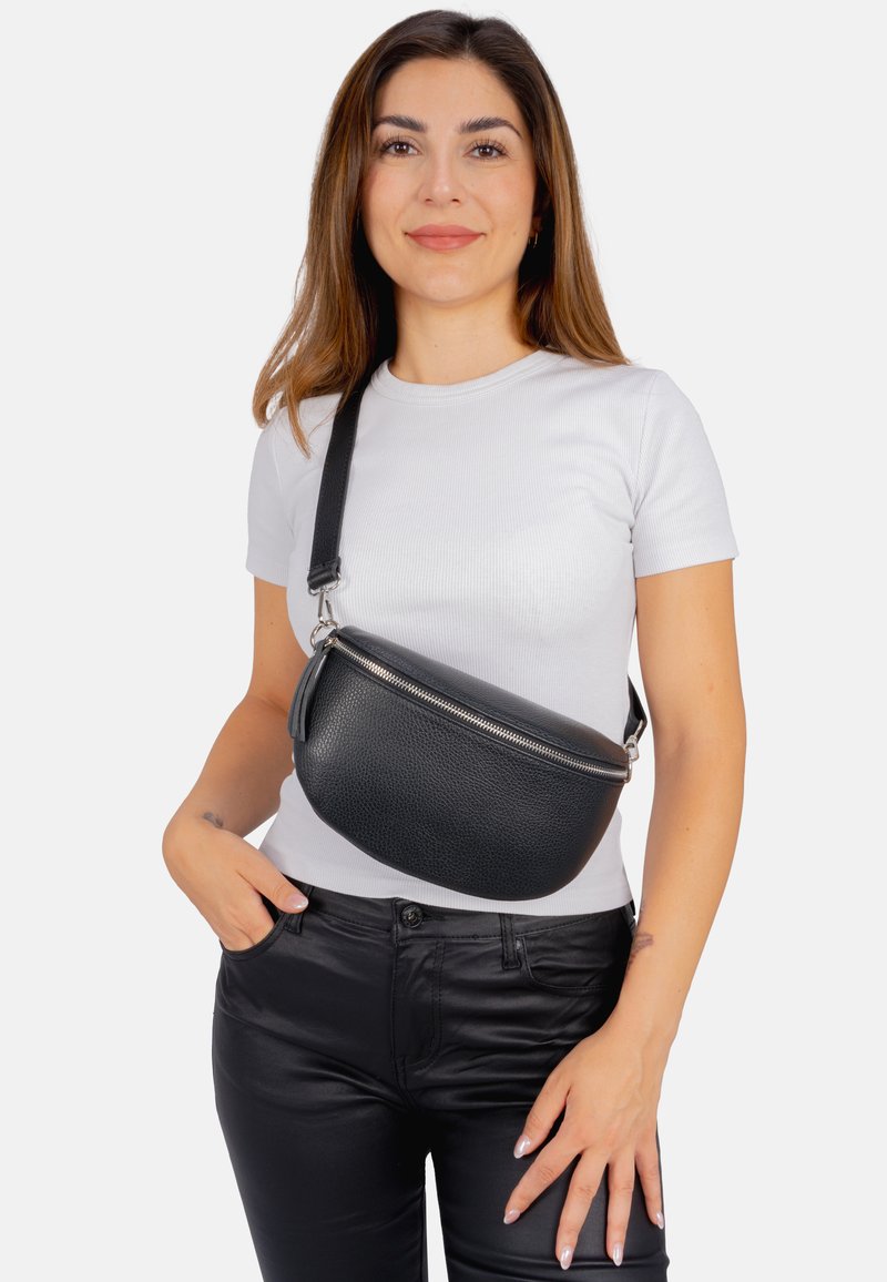 Schwarze Leder-Crossbody-Tasche mit strukturierter Oberfläche, runder Form, silbernem Reißverschluss und verstellbarem Trageriemen, getragen über einem weißen T-Shirt.
