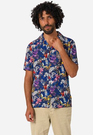 Man met krullend haar draagt een blauw overhemd met korte mouwen met een kleurrijk robotpatroon en beige broek tegen een effen achtergrond.