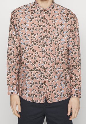 Homme portant une chemise à manches longues boutonnée avec un motif floral rose, marron et bleu et un pantalon sombre.