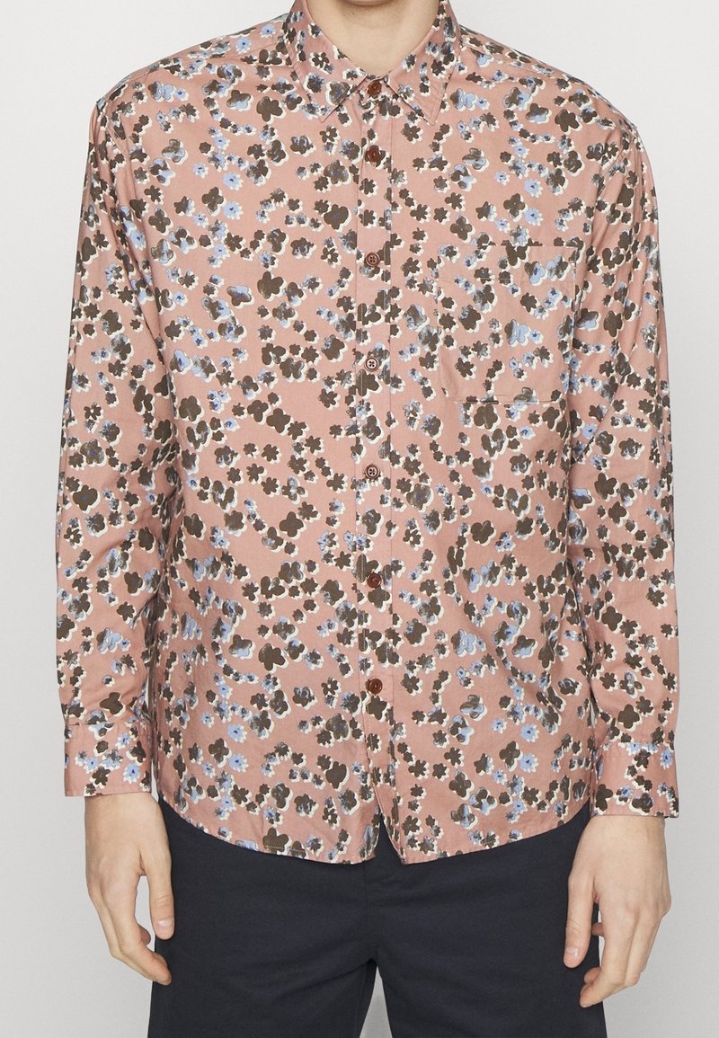 Homme portant une chemise à manches longues boutonnée avec un motif floral rose, marron et bleu et un pantalon sombre.