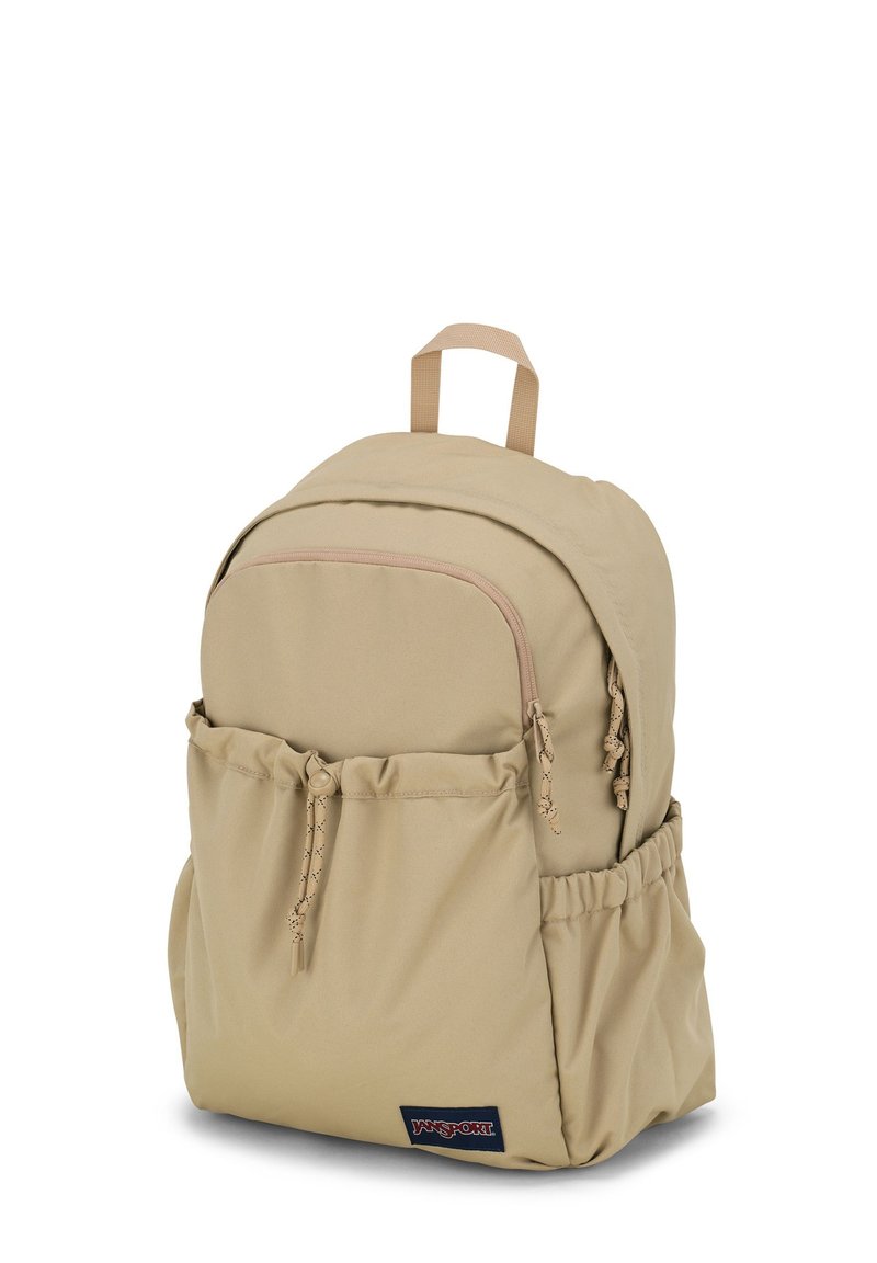 Sac à dos en tissu beige de forme arrondie, avec une poche zippée à l'avant, une poche latérale à cordon et une poignée de transport en haut. Badge logo sur le bas du devant.