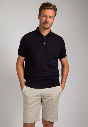 Zwart poloshirt met korte mouwen en een drieknops sluiting, gecombineerd met lichtbeige korte broek. De stof oogt zacht en aansluitend.