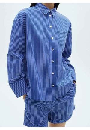 Žena v modro-bílé proužkované košili s dlouhým rukávem na knoflíky a sladěných šortkách, ruce v kapsách šortek, jednobarevné světlé pozadí.