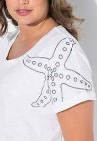 Camiseta de manga corta blanca con un diseño de estrella de mar negra en el hombro, adornos de costura y acentos circulares. Textura suave.
