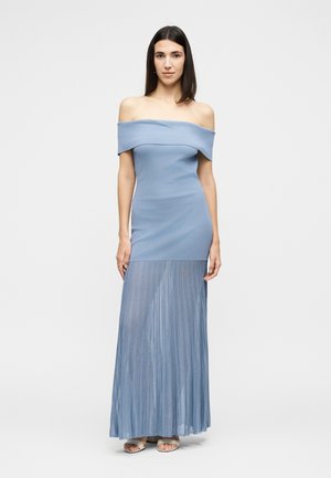 Γυναίκα στέκεται φορώντας ένα ανοιχτό μπλε, εφαρμοστό φόρεμα χωρίς ώμους με διάφανο πλισέ φούστα και μπεζ σανδάλια με ανοιχτά δάχτυλα σε μονόχρωμο φόντο.