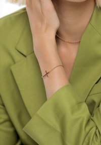 Roosakuldne kett- käevõru minimalistliku ristikujundusega, kantud rohelise jopega randmel. Tekstuur on sile ja elegantne.