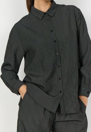 Femme portant une chemise noire à rayures fines boutonnée et un pantalon assorti, les mains dans les poches, légèrement de profil.