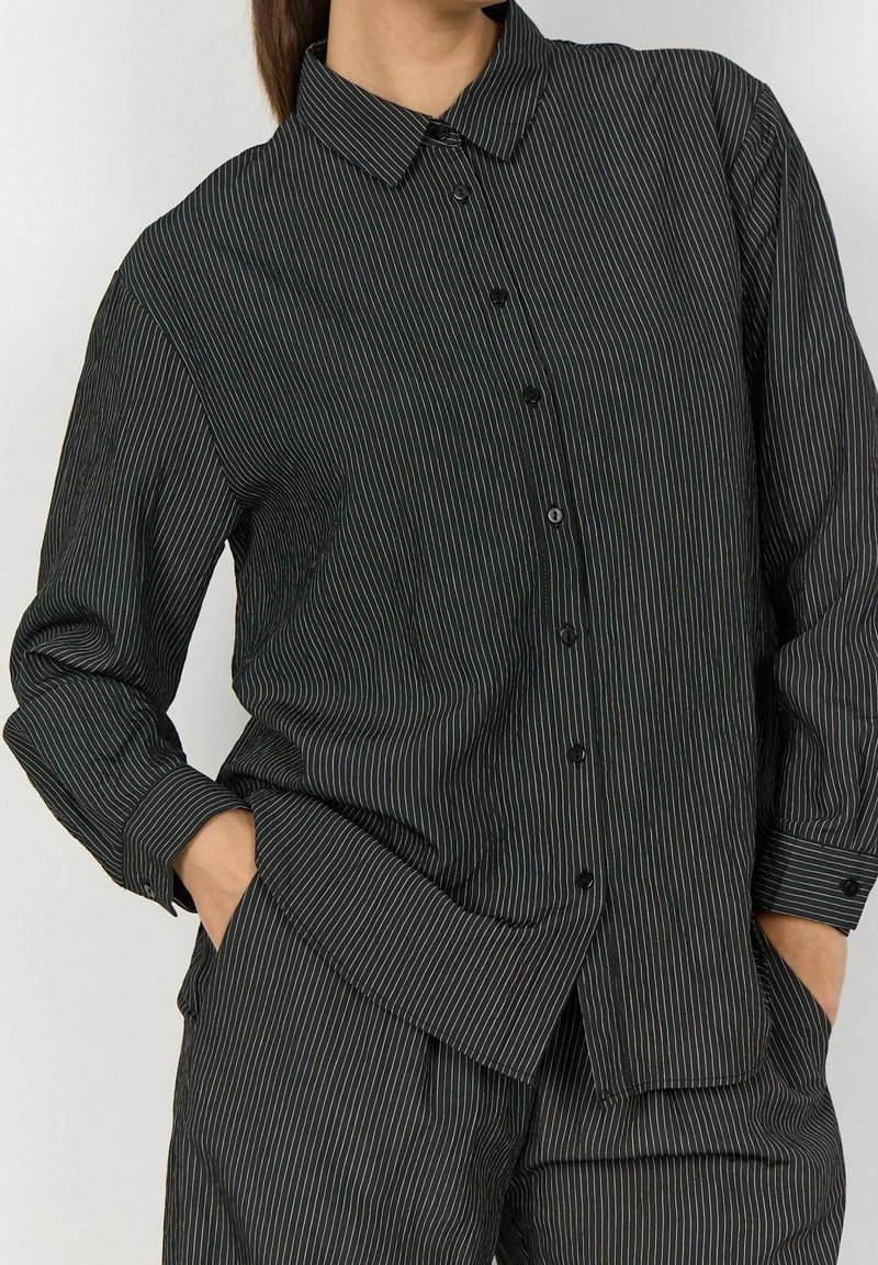Femme portant une chemise noire à rayures fines boutonnée et un pantalon assorti, les mains dans les poches, légèrement de profil.