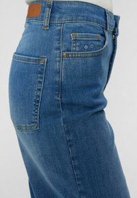 Denim kavbojke v srednje modri barvi z gladko teksturo. Imajo visok pas, pet žepov in kontrastne oranžne šive.