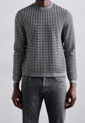 Hombre vistiendo un suéter de manga larga con patrón en blanco y negro, con cuello, puños y dobladillo grises, combinado con vaqueros grises desgastados.