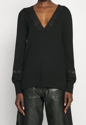 Jersey negro de manga larga con escote en V y ribete de encaje en el cuello y los puños, combinado con pantalones de cuero negro en una persona de piel oscura.
