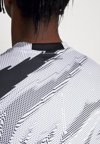 Camiseta deportiva en blanco y negro con un patrón de puntos y franjas negras marcadas, confeccionada en tejido que absorbe la humedad, con un escote redondo.