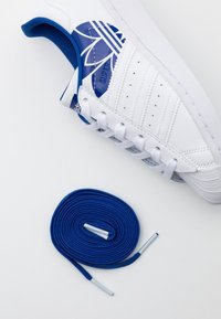 Valkoiset tennarit sinisillä yksityiskohdilla ja logolla, joissa on rei'itetyt sivut hengittävyyden lisäämiseksi ja litteät siniset kengännauhat muovikärjillä.