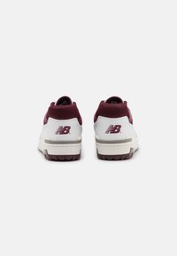 Pár bílých a burgundských tenisek New Balance zobrazených zezadu na jemně šedém pozadí.