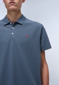 Grå polo skjorte laget av teksturert stoff, med en tre-knapps placket og et lite rødt og hvitt brodert emblem på venstre bryst.