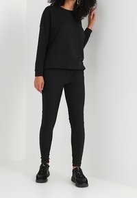 Svart sweatshirt med rund halsringning och nedhasade axlar, i kombination med svartvita randiga figurnära byxor. Svarta sneakers fulländar outfiten.