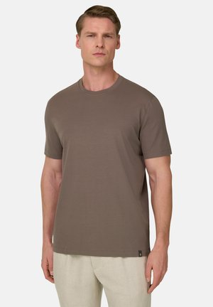 Homme portant un T-shirt marron uni à manches courtes et un pantalon beige clair, debout devant un fond gris clair uni.