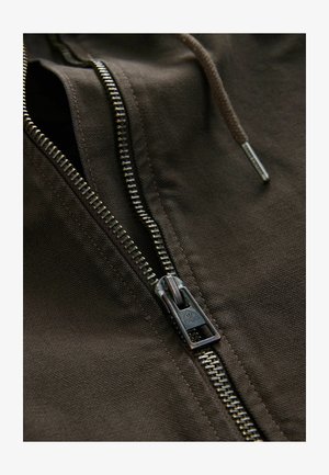 Brązowy zamek błyskawiczny z metalowym uchwytem, z blisko umiejscowionymi zębami i teksturowaną powierzchnią. Zamek jest częściowo otwarty.