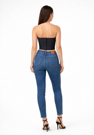 Donna in piedi, indossa un crop top scuro senza spalline, jeans skinny blu a vita alta e sandali neri con tacchi alti, di spalle su sfondo bianco.