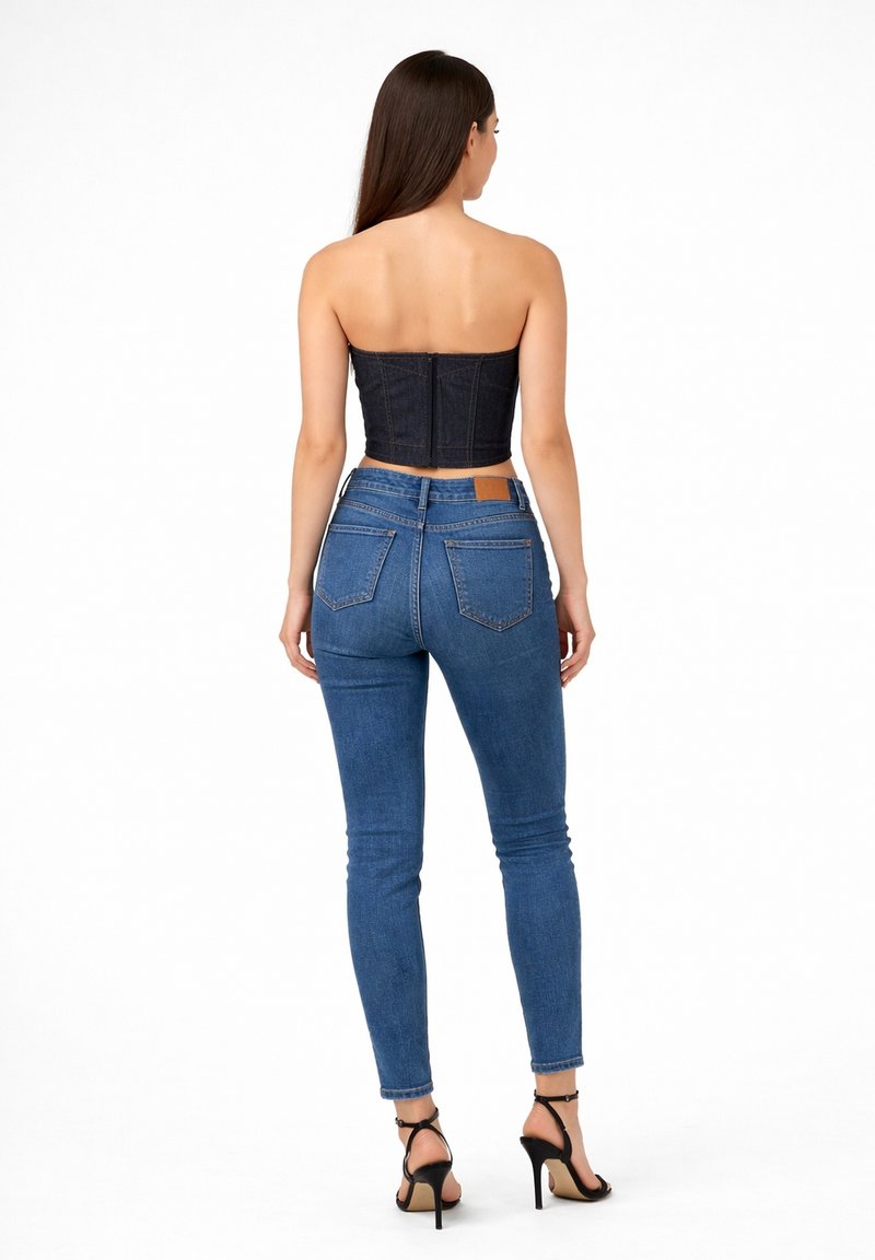 Mujer de pie, con top corto oscuro sin tirantes, vaqueros pitillo de talle alto azules y sandalias negras de tacón alto, de espaldas sobre fondo blanco.