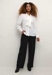 White long-sleeve blouse with a large bow detail at the collar, paired with loose-fitting black trousers and black heeled shoes.