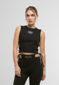 Top corto smanicato nero con lacci laterali e logo bianco. Abbinato a pantaloni neri con tagli e dettagli decorativi.