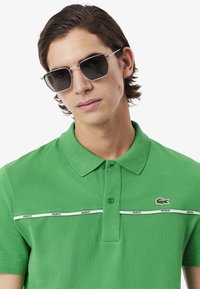 Groene polo met een gestructureerde stof, voorzien van een wit gestreept patroon en een klein logo. Model draagt een zonnebril met een metalen frame.