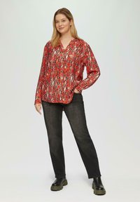 Langsleeve rode blouse met een slangenprint, gecombineerd met zwarte jeans en stevige zwarte laarzen. De blouse heeft een V-hals en een ontspannen pasvorm.
