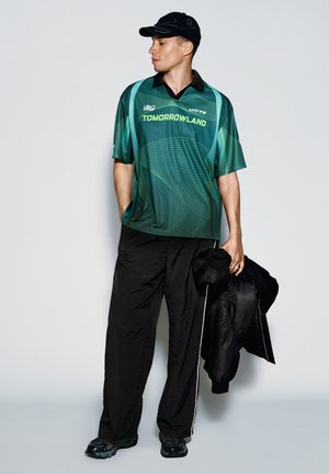 Man met zwarte pet, groen patroon Tomorrowland sportshirt, zwarte broek, een zwarte jas vasthoudend, staat tegen een effen achtergrond en kijkt naar links.